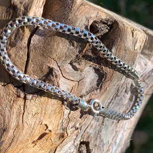 May include: A silver chain bracelet with a box chain design. The bracelet is lying on a piece of wood.