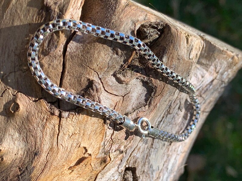 May include: A silver chain bracelet with a box chain design. The bracelet is lying on a piece of wood.