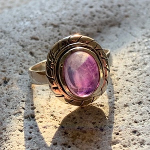Peut inclure: Une bague en argent ornée d'une pierre précieuse ovale de couleur violette. La pierre est sertie dans une lunette décorative et texturée. Le corps de la bague est en argent poli. La bague est présentée sur une surface texturée et claire.