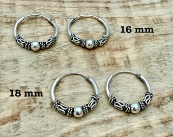 May include: Four silver hoop earrings arranged in two pairs. The top pair is labelled 16 mm, and the bottom pair is labelled 18 mm. Each hoop features a central smooth silver bead flanked by decorative twisted and scrolled metal patterns.