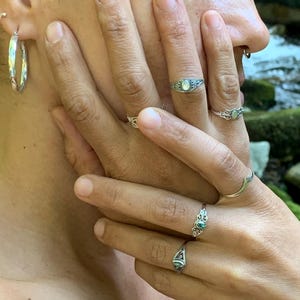 May include: Close-up of multiple silver rings with various designs, including a ring with a small, oval-shaped, light-colored stone. The image also shows a pair of silver hoop earrings and a nose ring.