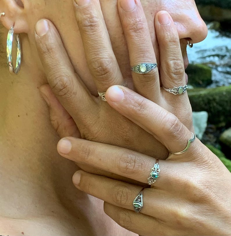 May include: Close-up of multiple silver rings with various designs, including a ring with a small, oval-shaped, light-colored stone. The image also shows a pair of silver hoop earrings and a nose ring.
