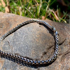 May include: A silver chain bracelet with a box chain design. The bracelet is lying on a grey rock.