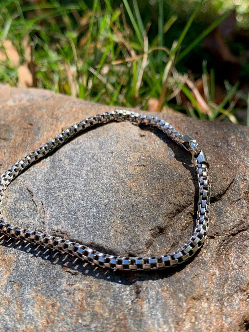 May include: A silver chain bracelet with a box chain design. The bracelet is lying on a grey rock.