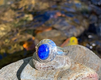 Può includere: Un anello d'argento con una pietra preziosa blu incastonata in una fascia d'argento decorativa. L'anello è appoggiato su una pila di pietre grigie.
