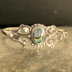 May include: A silver ring with an oval abalone shell center stone. The ring features ornate scrollwork on the sides and around the stone. The abalone shell displays iridescent colors of blue, green, and gold. The ring is set on a gray surface.