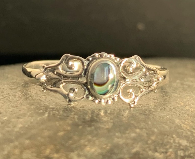 May include: A silver ring with an oval abalone shell center stone. The ring features ornate scrollwork on the sides and around the stone. The abalone shell displays iridescent colors of blue, green, and gold. The ring is set on a gray surface.