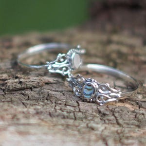 May include: Two silver rings with ornate filigree designs. One ring features a white stone, while the other has a blue and green abalone shell inlay. The rings are displayed on a textured wooden surface.