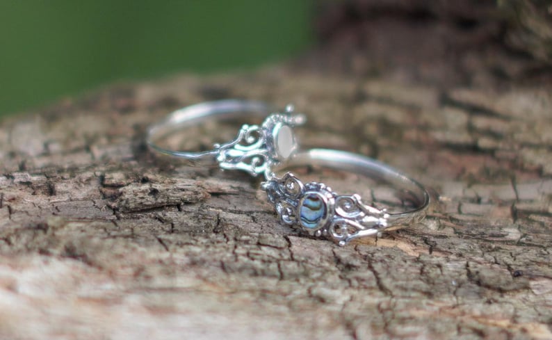May include: Two silver rings with ornate filigree designs. One ring features a white stone, while the other has a blue and green abalone shell inlay. The rings are displayed on a textured wooden surface.