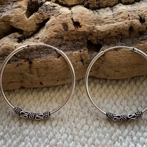 May include: A pair of silver hoop earrings with a decorative design on the hoop. The earrings are on a wooden surface.
