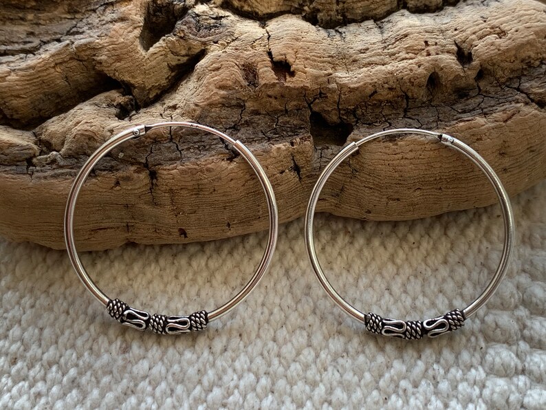 May include: A pair of silver hoop earrings with a decorative design on the hoop. The earrings are on a wooden surface.