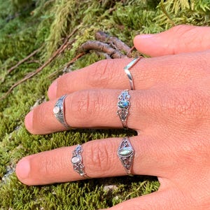 May include: A hand displays five silver rings. The rings feature various designs, including a chevron, ornate filigree, and oval-shaped stones in shades of white and blue-green. The background is a blurred view of green moss.