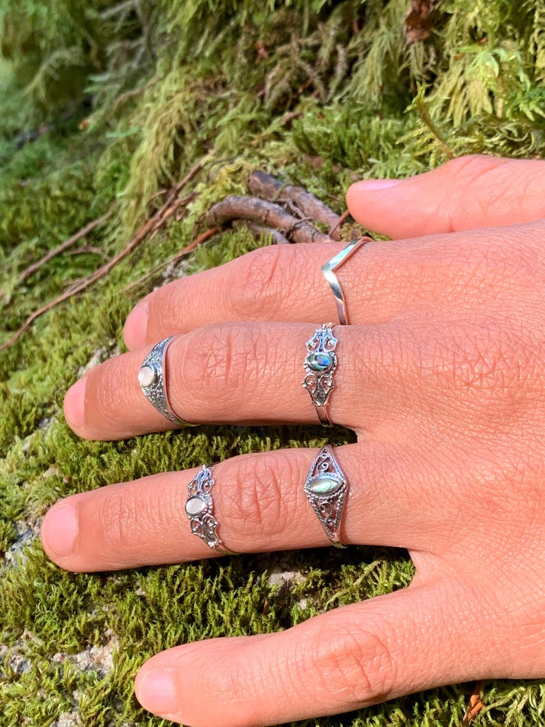 May include: A hand displays five silver rings. The rings feature various designs, including a chevron, ornate filigree, and oval-shaped stones in shades of white and blue-green. The background is a blurred view of green moss.