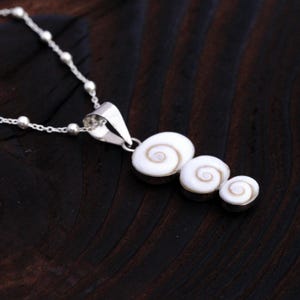 Könnte beinhalten: Eine Silberkette mit einem Anhänger aus drei unterschiedlich großen, weißen Shiva-Auge-Muscheln, die jeweils ein natürliches braunes Spiralmuster aufweisen. Der Anhänger ist an einer feinen Silberkette mit kleinen, gleichmäßigen Silberperlen befestigt.