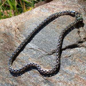 May include: A silver chain necklace with a lobster clasp closure. The chain is made of small, square links.