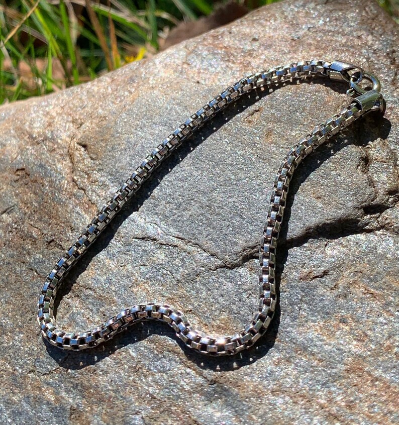 May include: A silver chain necklace with a lobster clasp closure. The chain is made of small, square links.