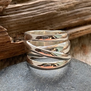 Pu&ograve; includere: Un anello d'argento con una fascia larga che presenta pi&ugrave; bande intrecciate. L'anello ha una finitura lucida e un design contemporaneo.