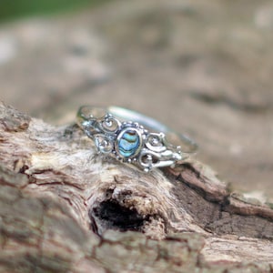 May include: A silver ring with an oval, iridescent abalone shell center stone. The ring has a decorative filigree design around the stone and along the band. The ring is displayed on a piece of textured, brown wood.