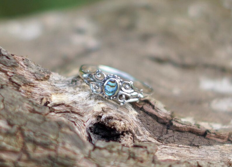 May include: A silver ring with an oval, iridescent abalone shell center stone. The ring has a decorative filigree design around the stone and along the band. The ring is displayed on a piece of textured, brown wood.