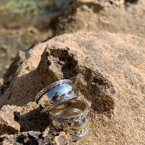 May include: Two silver rings with a braided design, stacked on top of each other, on a sandy surface.