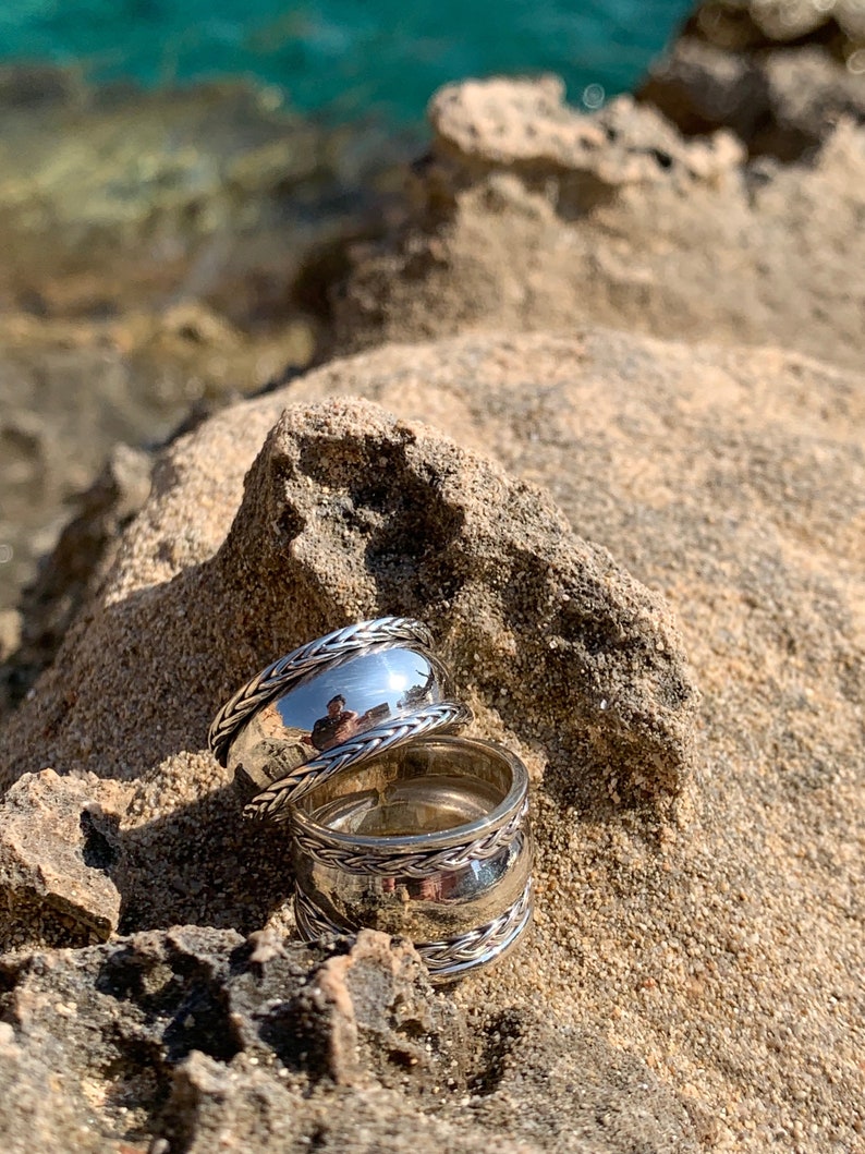 May include: Two silver rings with a braided design, stacked on top of each other, on a sandy surface.