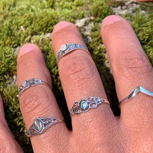 May include: Close-up of several silver rings with intricate designs and small gemstones. The rings are worn on fingers, showcasing their delicate details. The gemstones appear to be iridescent, reflecting various colors. The background is a soft green moss.