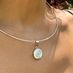 Peut inclure: Un collier argenté avec un pendentif. Le pendentif est une petite pierre blanche ronde avec un motif en spirale en fil d'argent. Le collier est porté autour du cou.