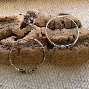 May include: A pair of silver hoop earrings with a decorative design on the hoop. The earrings are on a wooden surface.