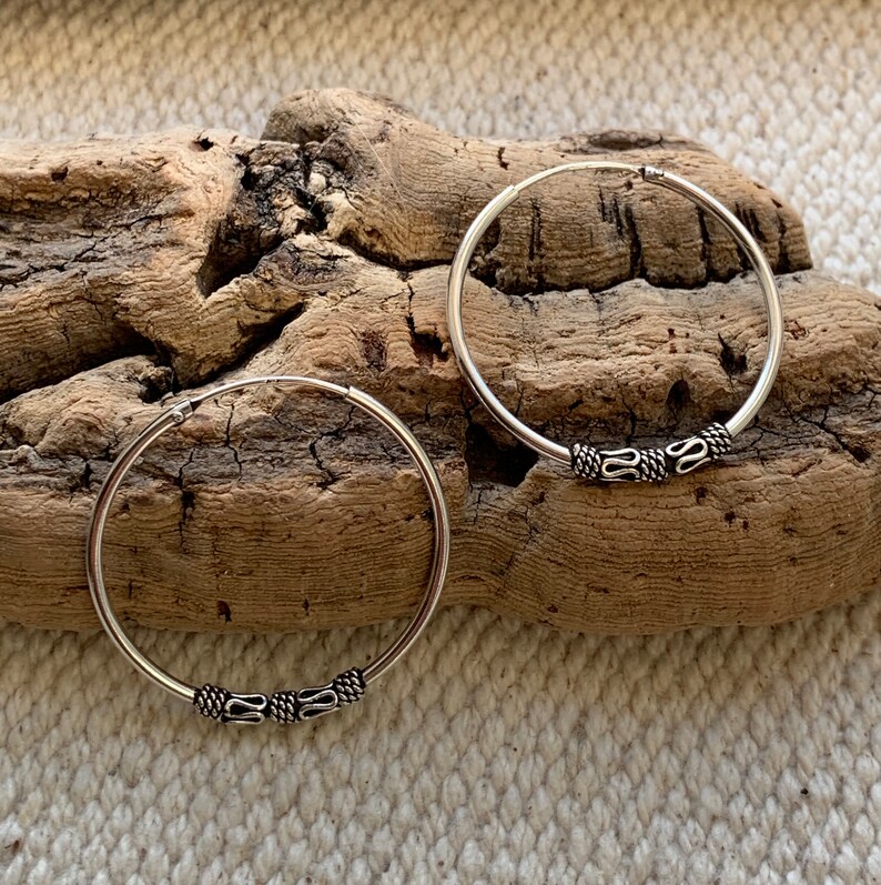 May include: A pair of silver hoop earrings with a decorative design on the hoop. The earrings are on a wooden surface.