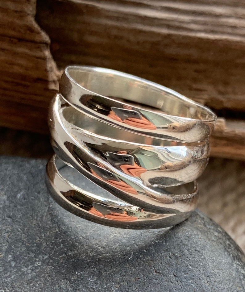 Pu&ograve; includere: Un anello d'argento con una fascia larga composta da tre pezzi curvi sovrapposti. L'anello &egrave; lucido e brillante.