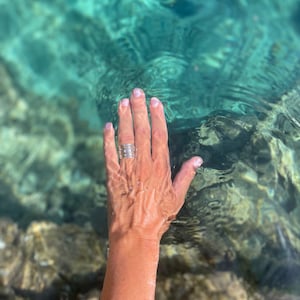 K&ouml;nnte beinhalten: Eine Hand mit einem silbernen Ring mit mehreren B&auml;ndern greift in klares t&uuml;rkisfarbenes Wasser.
