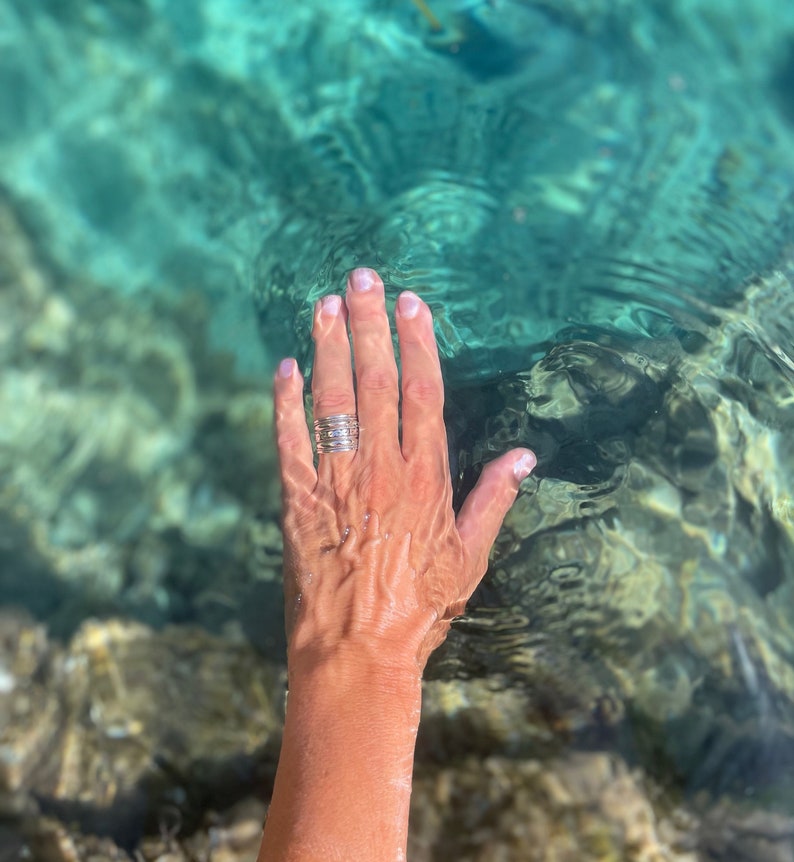 K&ouml;nnte beinhalten: Eine Hand mit einem silbernen Ring mit mehreren B&auml;ndern greift in klares t&uuml;rkisfarbenes Wasser.