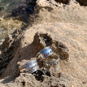 May include: Three silver rings with a braided design. The rings are stacked on top of each other and are sitting on a rocky surface.