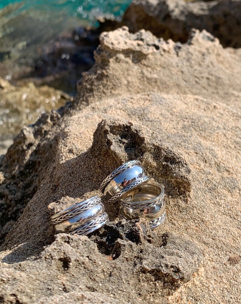 May include: Three silver rings with a braided design. The rings are stacked on top of each other and are sitting on a rocky surface.