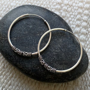 May include: A pair of silver hoop earrings with decorative accents. The hoops are smooth and have a shiny finish. The accents are made of silver and have a textured design.