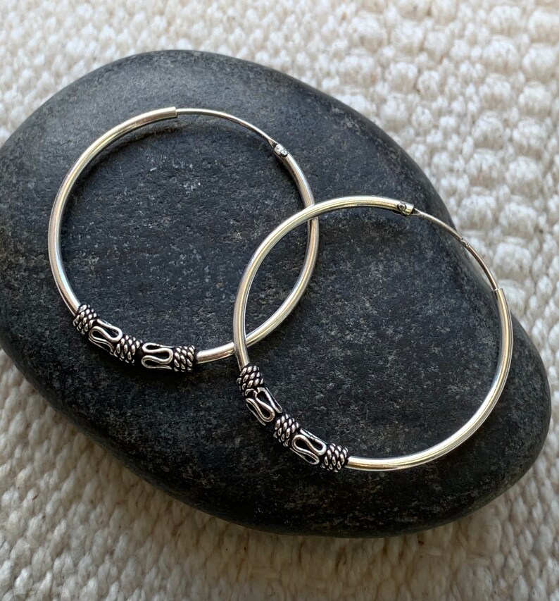 May include: A pair of silver hoop earrings with decorative accents. The hoops are smooth and have a shiny finish. The accents are made of silver and have a textured design.