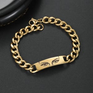 May include: A gold-toned metal chain link bracelet featuring a central rectangular identification plate. The plate is engraved with a detailed black graphic illustration of a pair of human eyes with defined eyebrows and eyelashes.