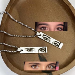 May include: Two silver-toned necklaces with rectangular pendants featuring eye illustrations. The pendants hang from a chain with a textured pattern. The necklaces are displayed on a wooden tray.