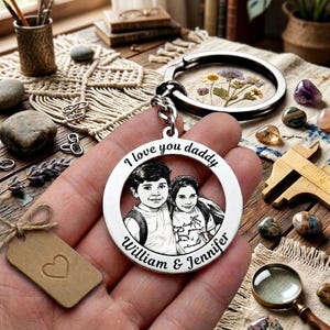 May include: A silver keychain with a circular pendant featuring a sketch of two children and the words "I love you daddy," "William & Jennifer." The keychain is held in a hand, with a tag and other items in the background.