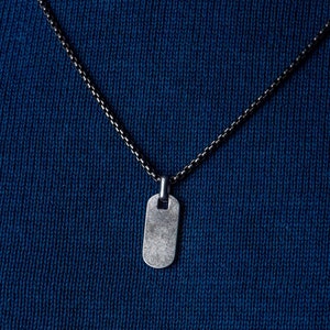 May include: A silver dog tag pendant on a silver chain necklace against a dark blue background.