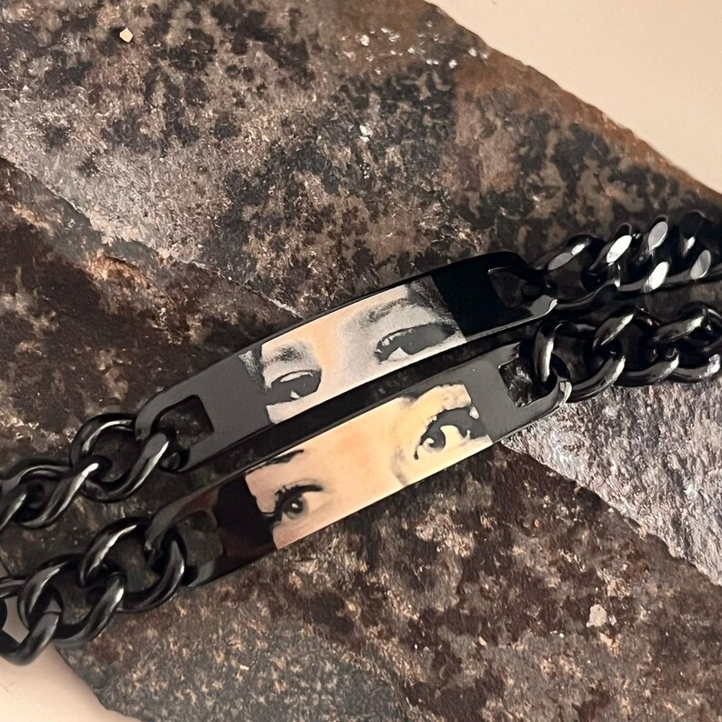 May include: Two black metal chain bracelets featuring rectangular plates. Each plate displays a detailed, high-contrast photographic print of a pair of human eyes. The bracelets are constructed with thick, interlocking links in a dark finish.