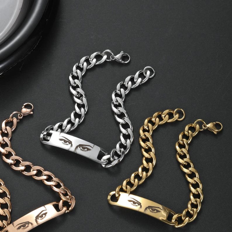 May include: Three metal chain bracelets in silver, gold, and rose gold tones. Each bracelet features a flat rectangular center plate engraved with a detailed pair of human eyes. The chains consist of interlocking oval links with a lobster clasp.