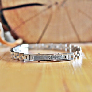 May include: A silver bracelet with a brushed metal finish. The bracelet has a rectangular plate with two fingerprints and the letters "B & H" engraved on it.