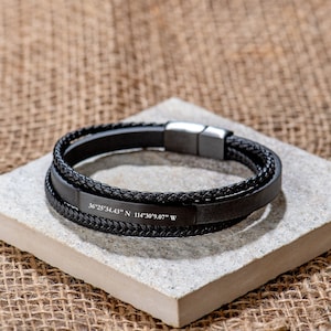 Puede incluir: Pulsera de cuero negro con tres capas de cuero trenzado y liso. La pulsera tiene un cierre plateado y coordenadas grabadas: 36°25'34.43" N 114°30'9.07" W.