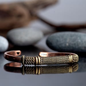 Op de afbeelding: Een koperen manchetarmband met een geweven goudkleurig middengedeelte. De armband heeft een klein zwart hartsymbool en de woorden "I love you" gegraveerd. De armband wordt gepresenteerd op een reflecterend oppervlak.