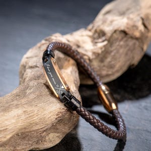 May include: Brown braided leather bracelet with a black and gold clasp. The clasp features the inscription "I love you more" and a compass design. The bracelet is shown on a piece of driftwood.