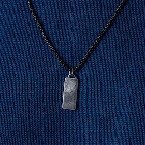 May include: A silver rectangular pendant hangs from a black chain necklace on a blue knitted fabric background.