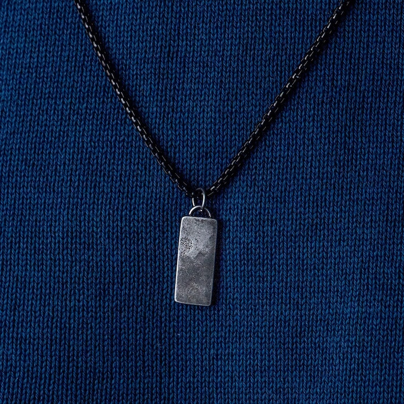 May include: A silver rectangular pendant hangs from a black chain necklace on a blue knitted fabric background.
