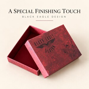 May include: A rectangular red gift box with a textured finish, featuring a black eagle logo and floral line art. The lid is partially open, revealing a solid red interior. The text A SPECIAL FINISHING TOUCH BLACK EAGLE DESIGN is visible.