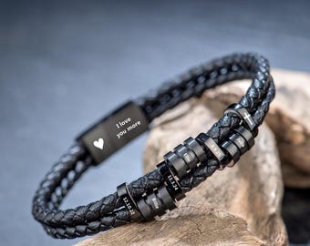May include: Black braided leather bracelet with personalised beads. The bracelet features the text "I love you more" and a heart symbol. The beads are engraved with names and dates. The bracelet is displayed on a piece of wood.